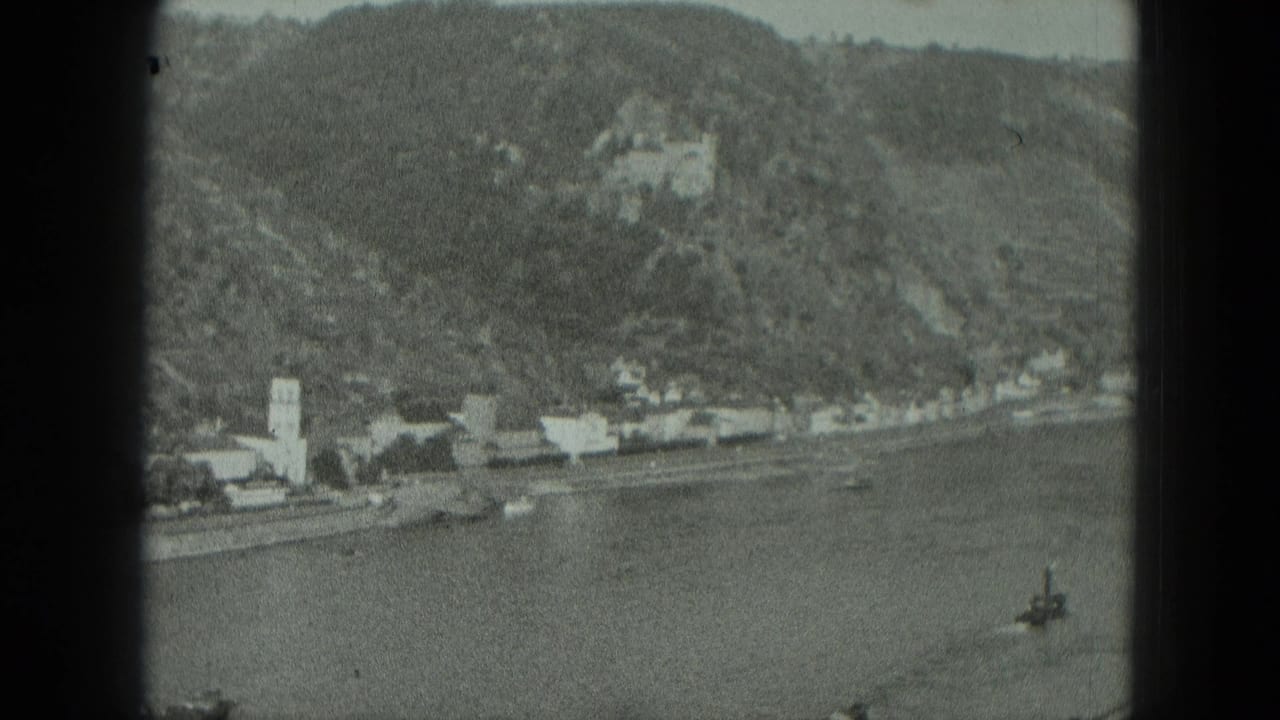 1951 West Germany 8mm frame showing a river, shoreline buildings, church steeple, wooded hillside, and castle above the village.