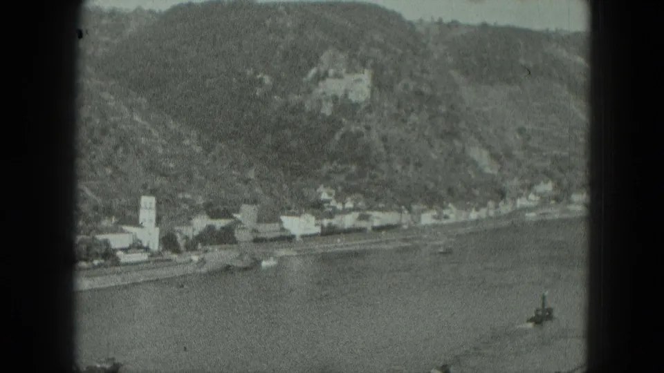 1951 West Germany 8mm frame showing a river, shoreline buildings, church steeple, wooded hillside, and castle above the village.