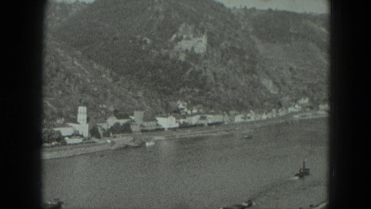 Wide 1951 West Germany view of a riverfront village, church steeple, boats on the water, and a castle on the wooded hillside.