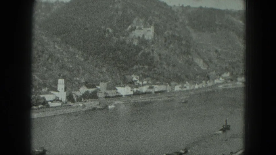 Wide 1951 West Germany view of a riverfront village, church steeple, boats on the water, and a castle on the wooded hillside.