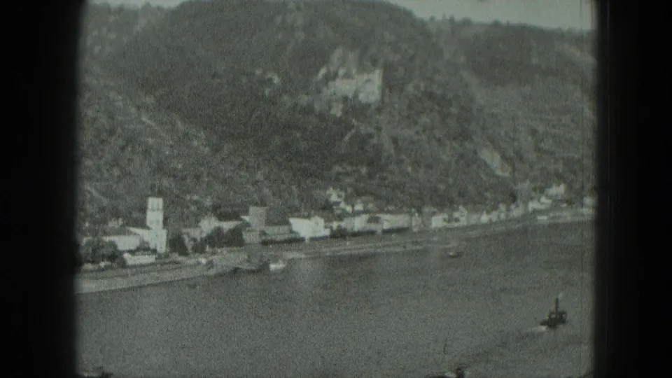 Black-and-white 8mm frame of a West German river village in 1951, with shoreline buildings, church steeple, hillside, and castle visible.