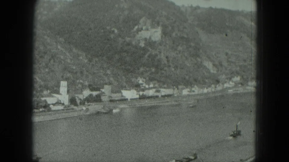 1951 West Germany home-movie frame showing a river, clustered shoreline buildings, a church tower, and a distant castle on the hill.