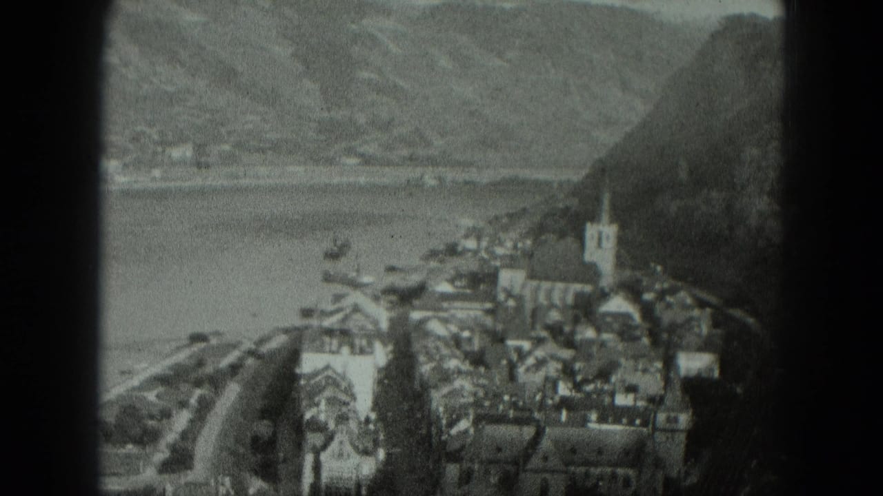 Aerial view of a clustered waterfront village with a church tower, bay, boats, and hills.