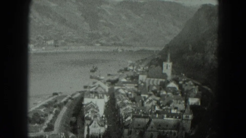 1951 West Germany 8mm aerial view showing a village beside calm water, with rooftops, a church steeple, boats, and surrounding hills.