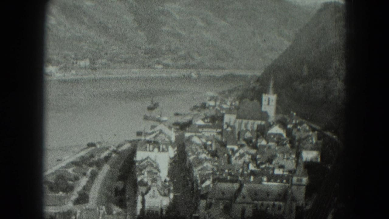 Black-and-white 8mm frame of a West German waterside village, with a prominent church steeple, dense roofs, small boats, and mountain slopes.