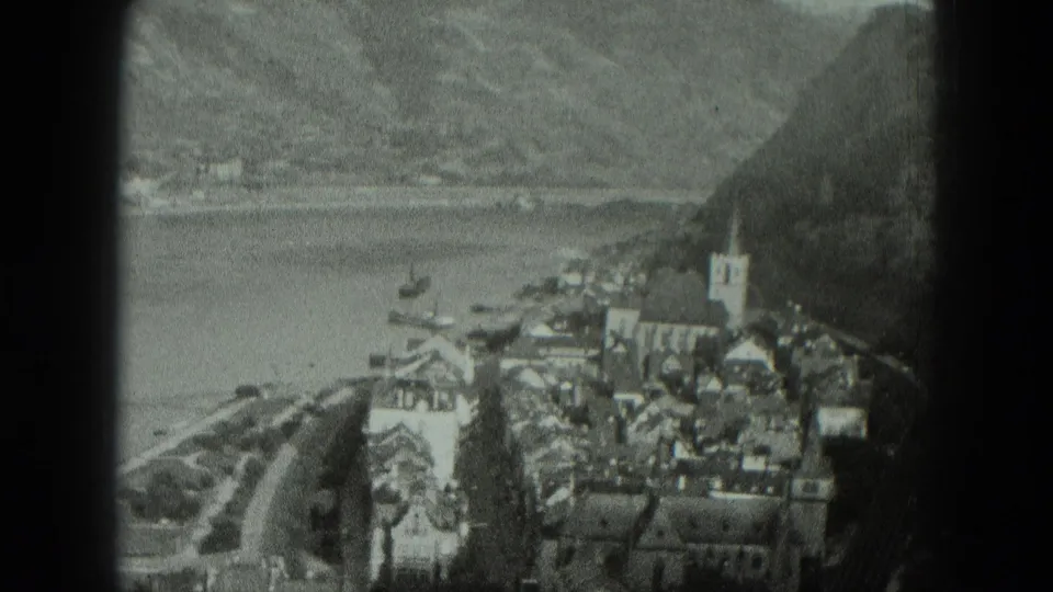 Black-and-white 8mm frame of a West German waterside village, with a prominent church steeple, dense roofs, small boats, and mountain slopes.