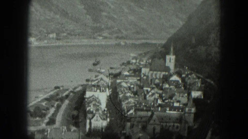 1951 black-and-white home-movie frame looking down on a West German village beside water, including a church tower, boats, road, and hills.