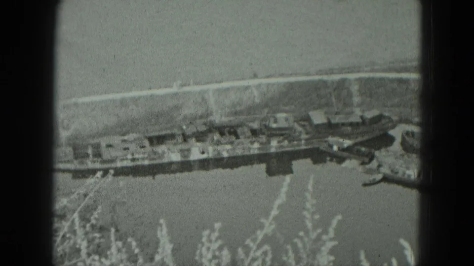 Black-and-white 8mm frame showing a West German harbor or pier area from above, with waterside buildings, shoreline, road, and vegetation.