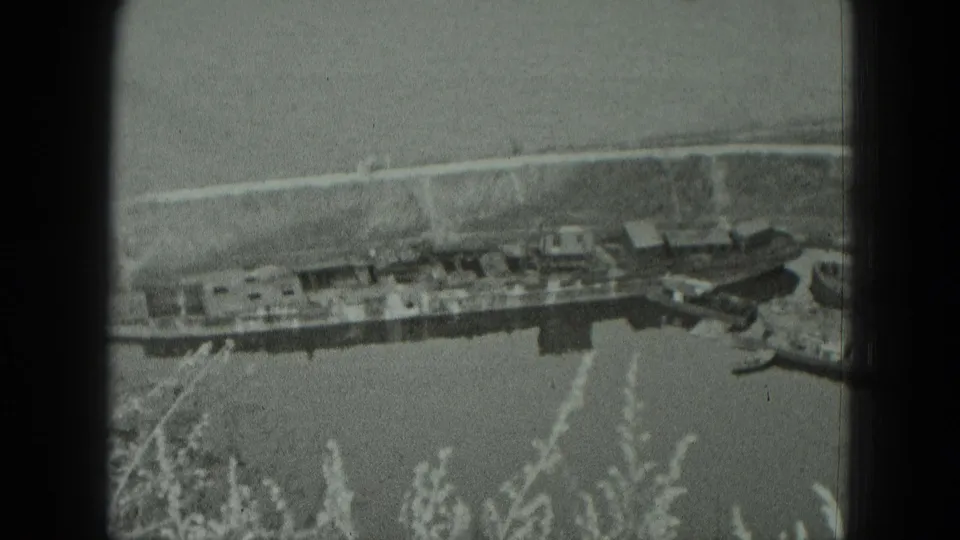 1951 West Germany 8mm frame showing moored boats beside a low riverbank, with reeds soft in the foreground.