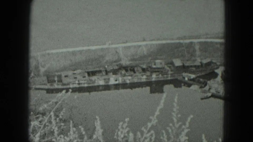 1951 West Germany riverside view with a long moored vessel, smaller boats, a pale bank, and soft film grain.