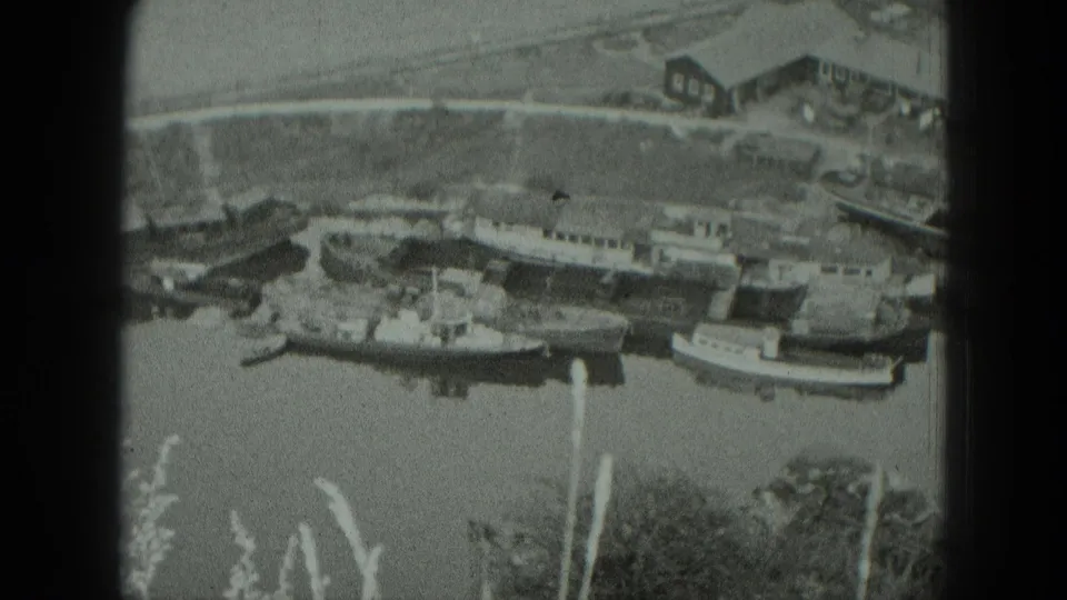 1951 West Germany home-movie frame showing clustered boats, modest shore buildings, reeds, and a calm river surface.
