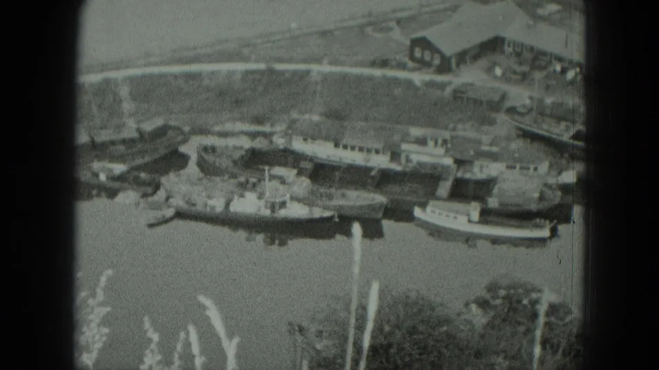 1951 West Germany 8mm view of boats tied along the riverbank below houses or riverside buildings, framed by reeds.