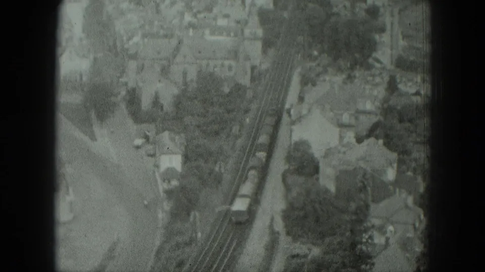 1951 West Germany home-movie frame showing a train on tracks through a residential area with roads, rooftops, and trees.
