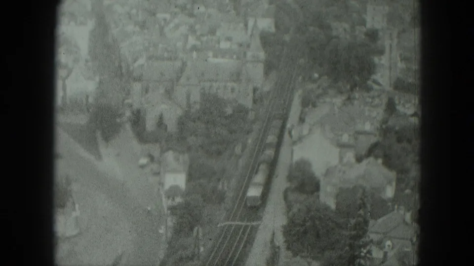 Grainy 1951 West Germany frame with a train traveling along tracks near houses, trees, and a road under cloudy daylight.