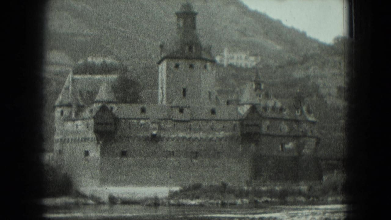 Fortified riverside structure with towers, wall, riverbank, and hillside behind it.