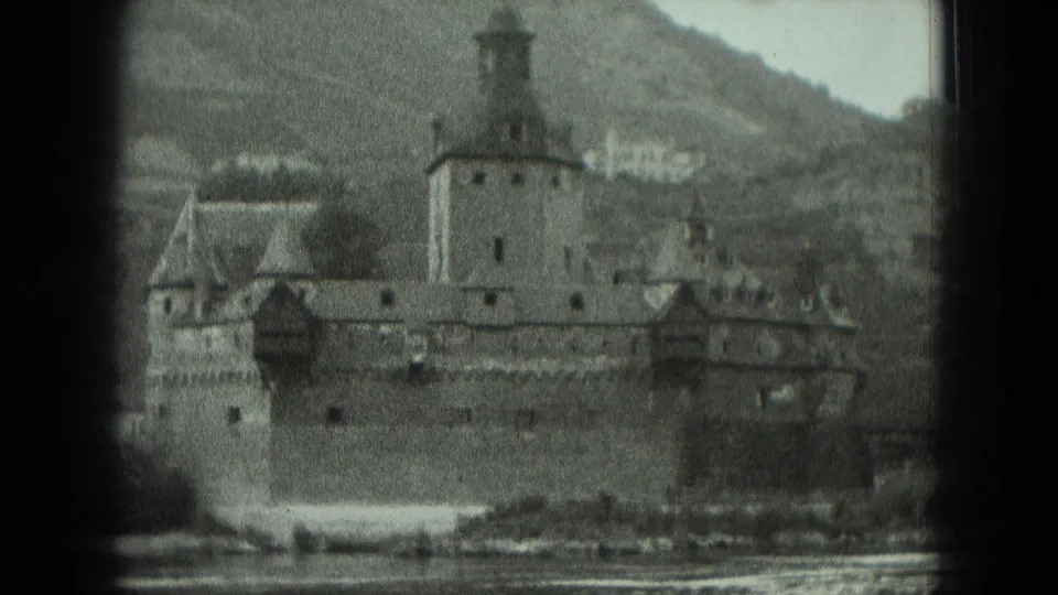 1951 Germany home-movie frame showing a large riverside fortress with towers, stone walls, water in front, and a wooded hillside behind.