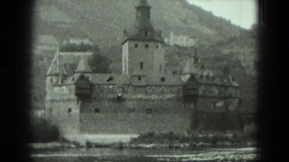 1951 Germany frame of a riverside fortress from across the water, showing towered stone walls, a shoreline, and hills behind the site.