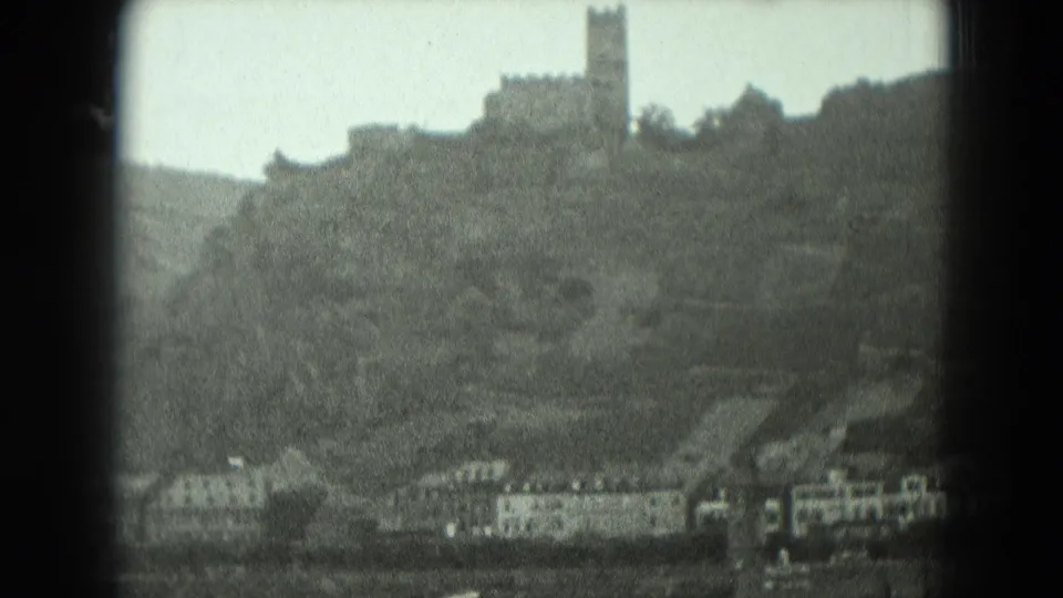 Soft black-and-white view of a German hillside castle and tower above riverside structures in 1951 home-movie footage.