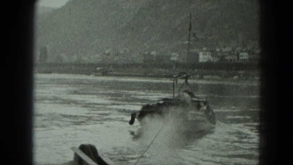 Black-and-white 8mm frame from a river boat, with foreground rigging and distant German shoreline buildings.
