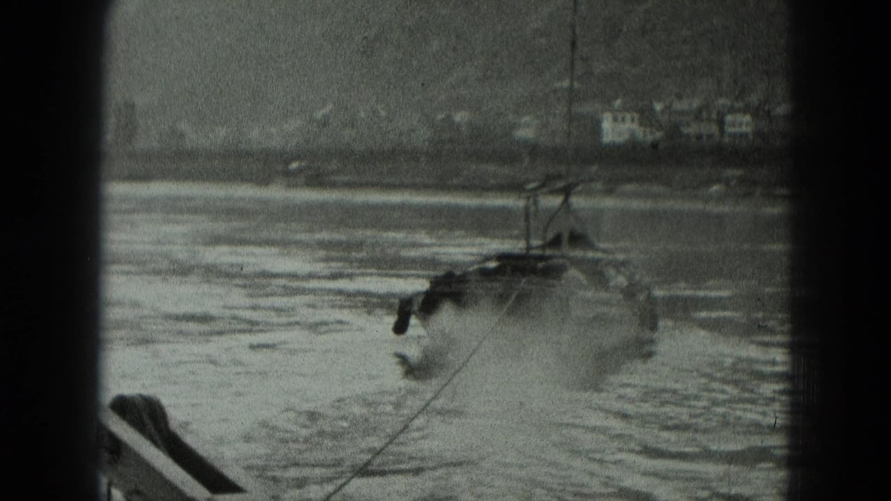 Military-style boat moving across a river with hills and buildings in the background.