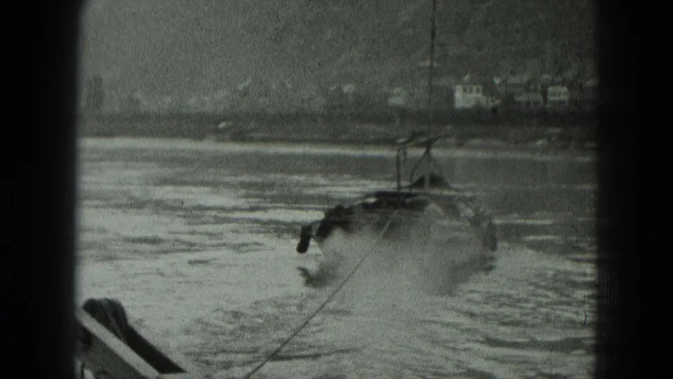 Black-and-white 1951 8mm frame of a military-style boat crossing a river in Germany, with hillside buildings beyond the water.