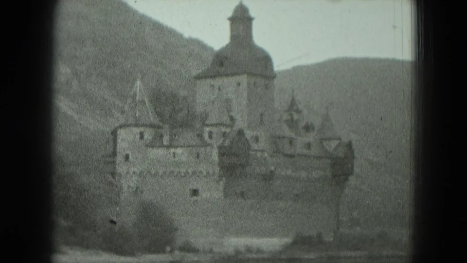 Black-and-white 1951 8mm frame showing a German riverside castle-like building with towers, turrets, and hills behind it.