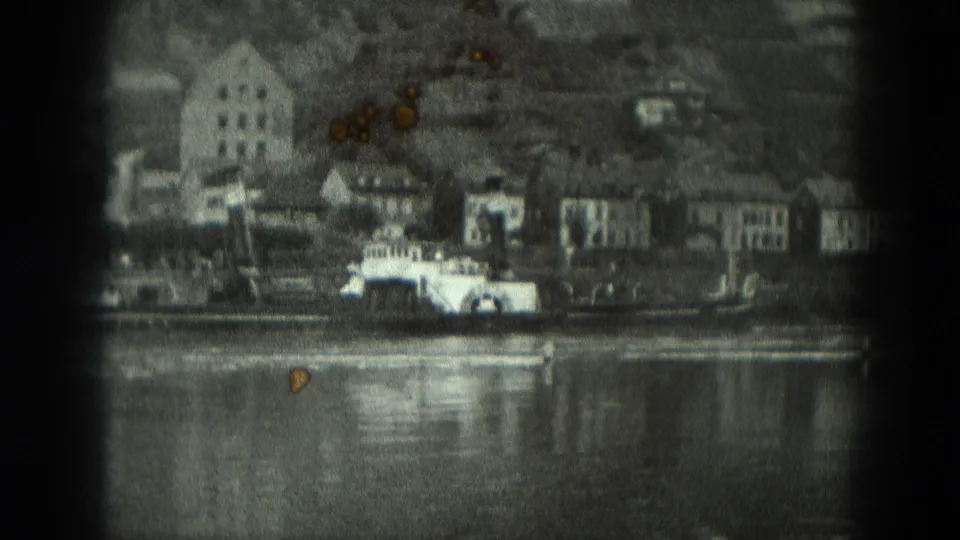 Grainy 1951 West Germany river-town frame showing waterfront houses, a paddlewheel-style vessel, hillside terrain, and water reflections.