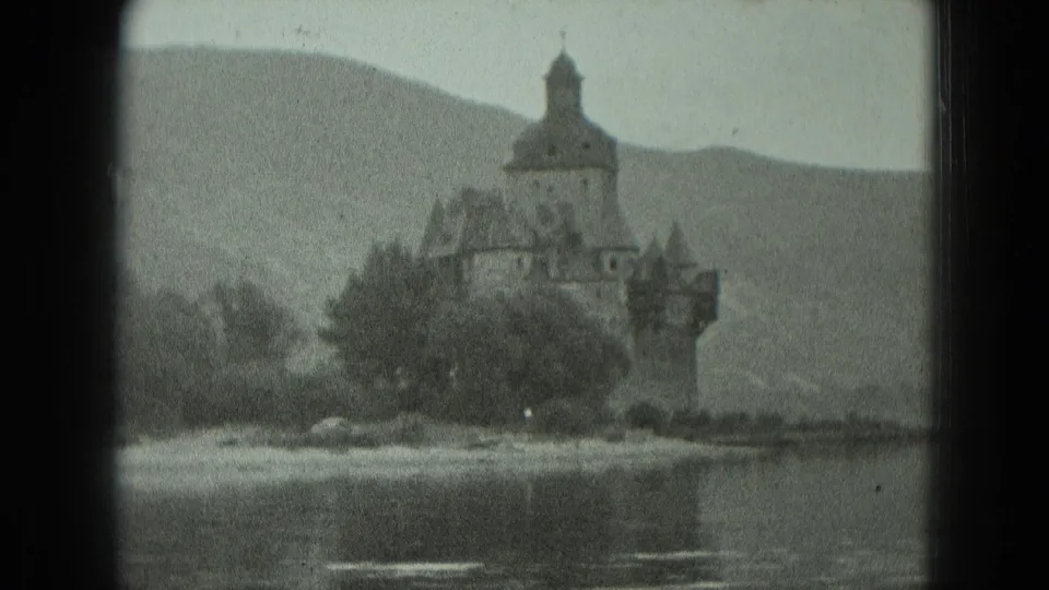 1951 West Germany home-movie frame of a castle-like waterfront building reflected near calm water with hills beyond.
