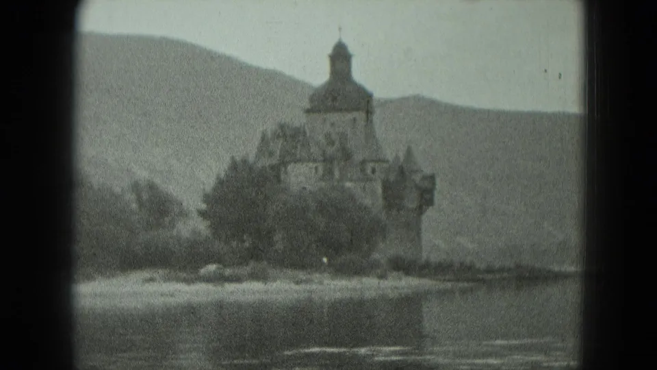 Soft monochrome 8mm view of a waterfront castle-like structure in Germany, bordered by trees and backed by hazy hills.
