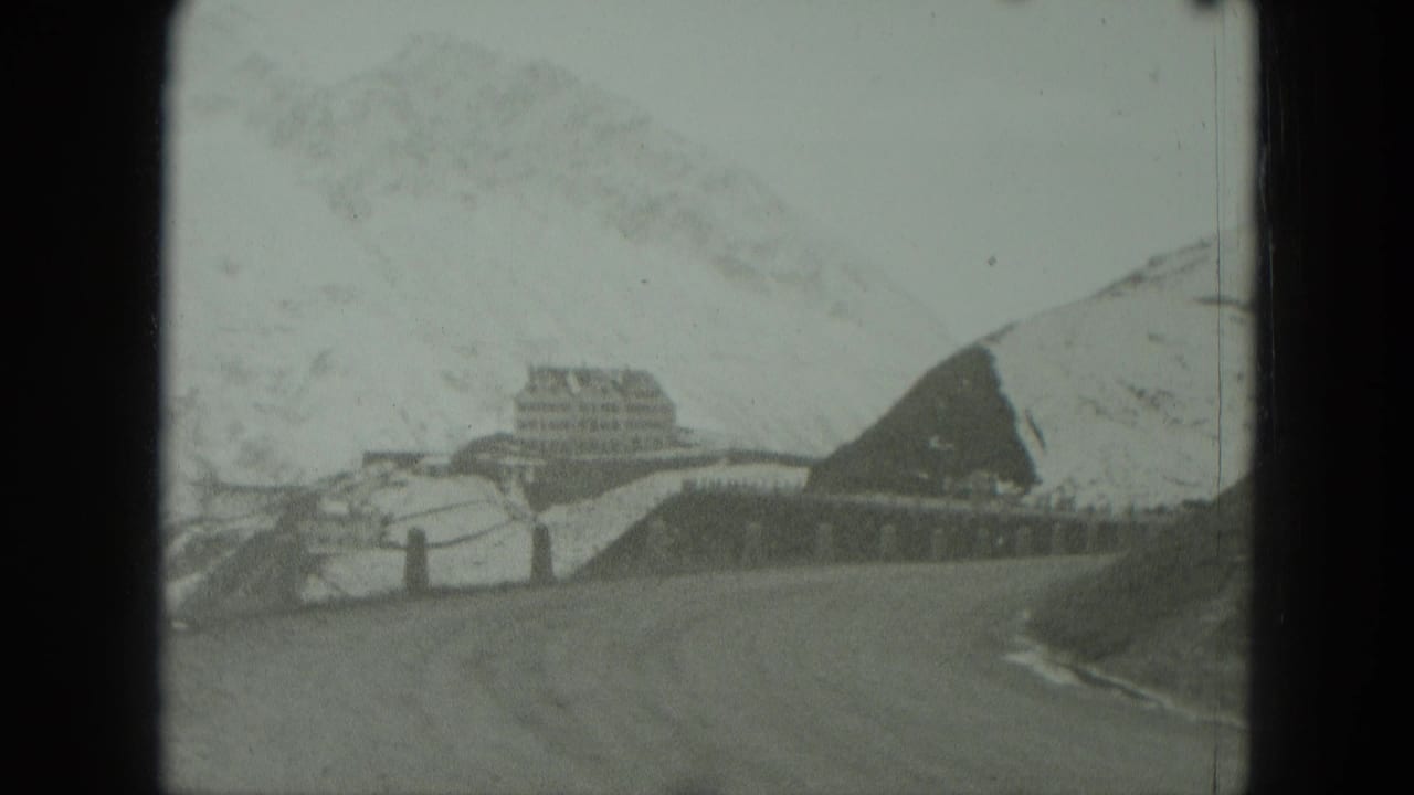 Snowy mountain road leading toward a large distant building under a hazy sky.
