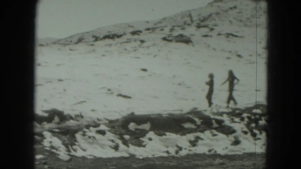 Two distant people walk across snowy alpine ground in Germany, with rocky patches and a broad white slope behind them.
