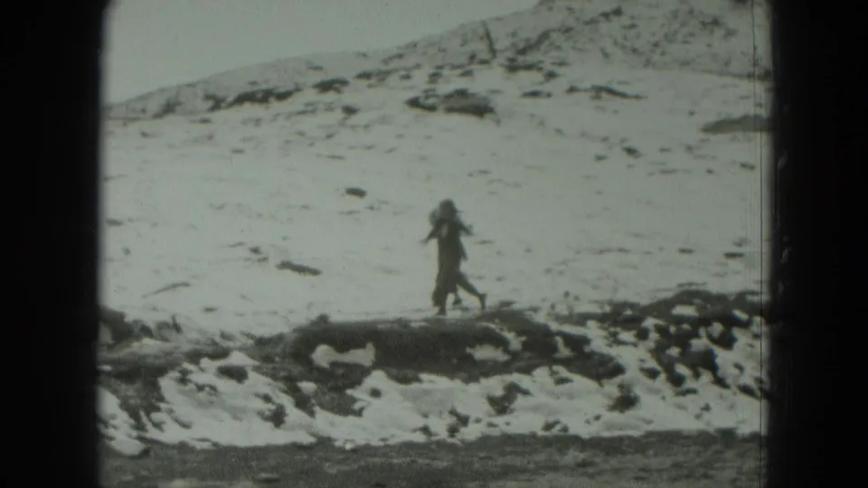A distant walker crosses rocky snow in a German mountain landscape, seen in grainy 8mm home-movie footage.