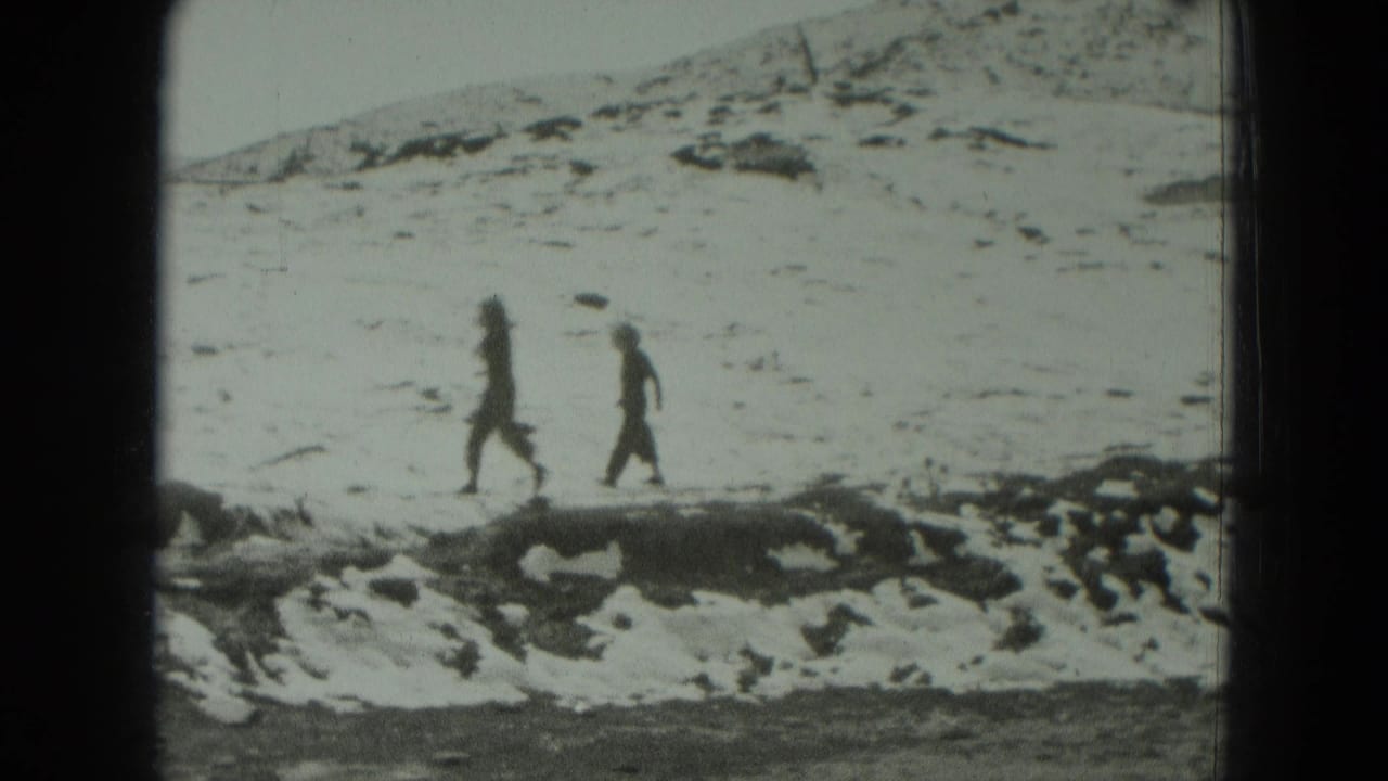 Two dark-clad figures walk across a snowy hillside with rocks in the foreground.