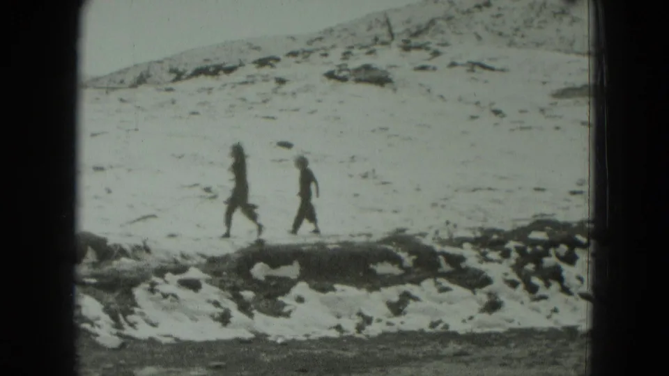 In 1951 West Germany home-movie footage, two distant figures walk across a snow-covered hillside under a cloudy sky.