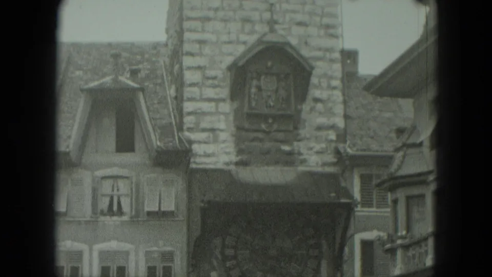 A stone clocktower with a small clock or emblem is framed among steep-roofed European buildings in 1951 Germany footage.