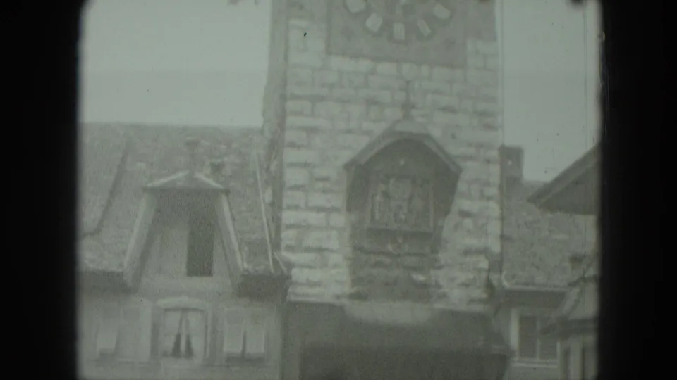 1951 West Germany 8mm frame showing a stone clock tower with a large clock face and cuckoo-clock feature over nearby roofs.