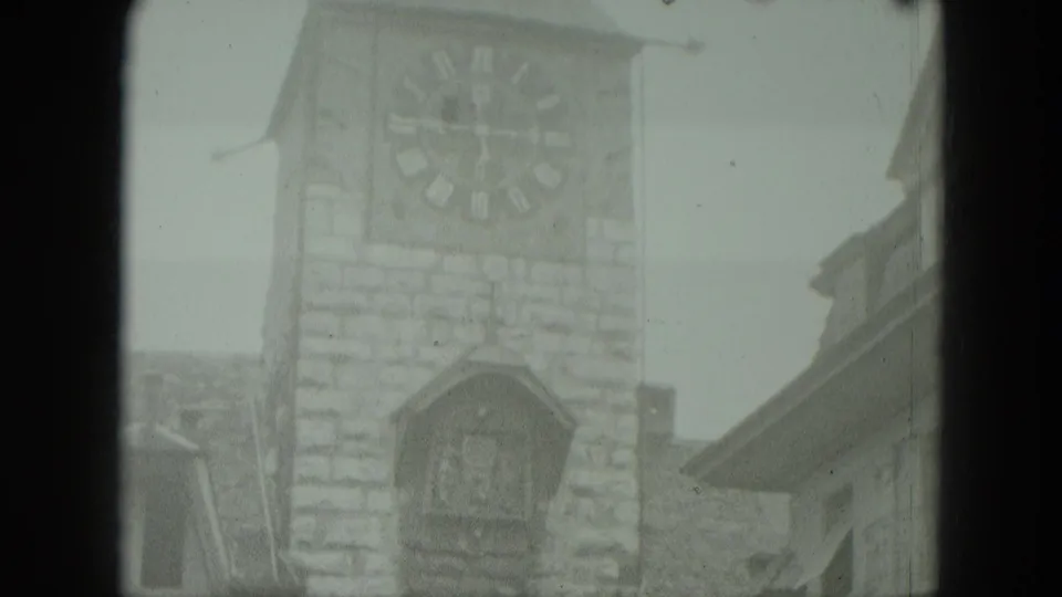 Soft 8mm view of a West German clock tower with a large clock face, cuckoo-clock detail, and adjoining rooftops.