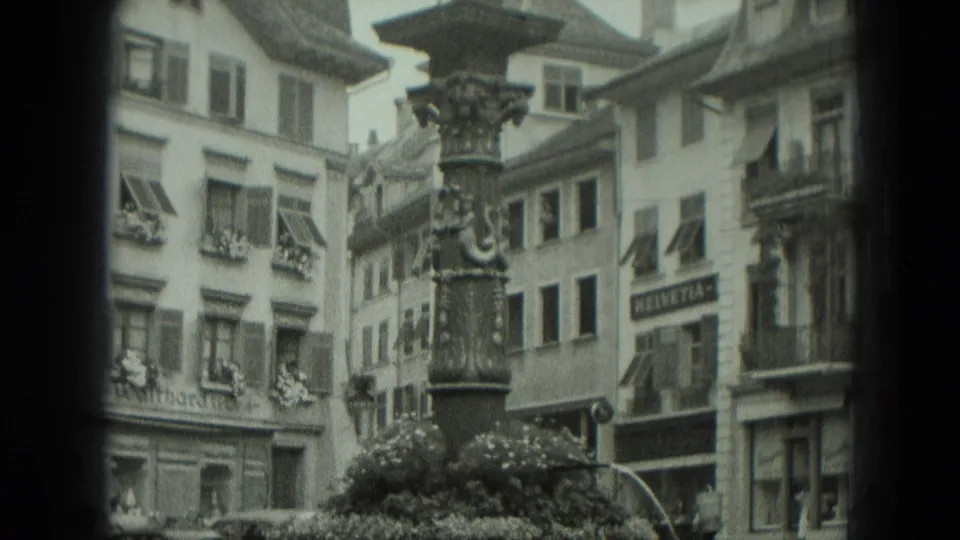 1951 West Germany frame with an ornate fountain column, shuttered facades, flower boxes, shopfronts, and a visible HELVETIA sign.