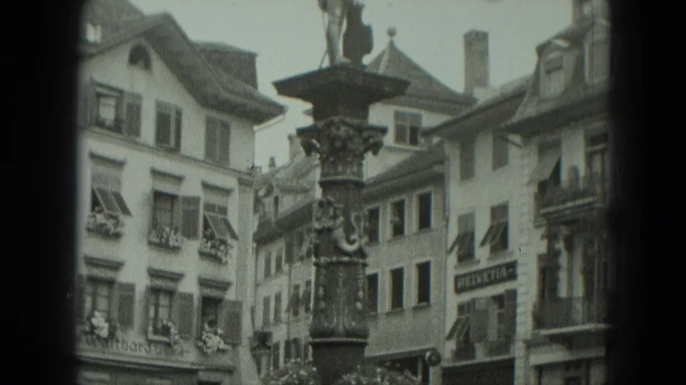 Black-and-white 8mm frame from 1951 West Germany showing the ornate fountain column before shuttered town-square buildings.