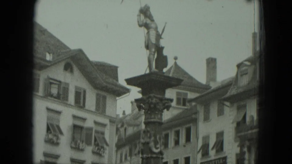 1951 West Germany 8mm frame showing a flag-bearing statue on a column with gabled buildings and shuttered windows behind it.