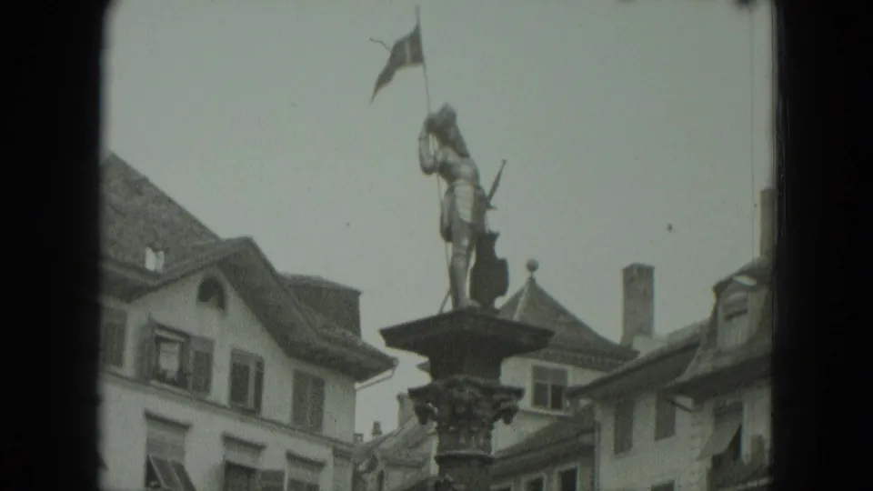 1951 West Germany frame with the column statue shifted right, backed by steep roofs, shutters, and a flag near the roofline.