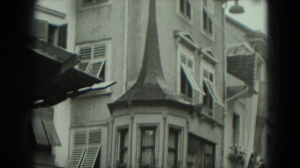 1951 Germany 8mm frame looking up at a facade with a pointed roof detail, bay window, louvered shutters, and nearby street fixtures.