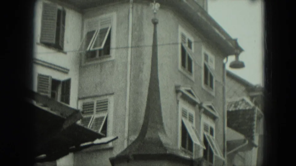 1951 Germany 8mm frame showing the upper stories of an older building with a pointed roof form, shutters, windows, and a hanging street lamp.