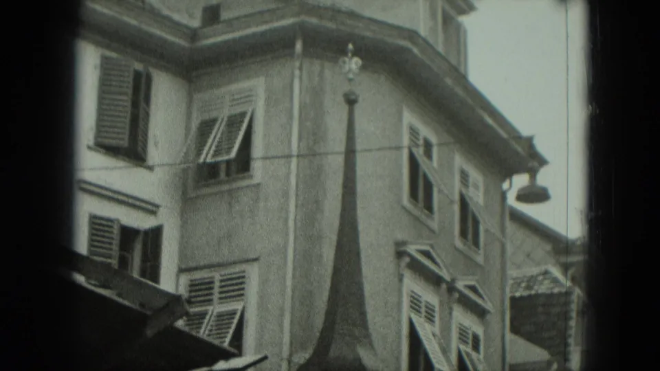 1951 Germany home-movie frame showing shuttered stucco buildings, a steep roofline and street lamp partially obscured by film borders.