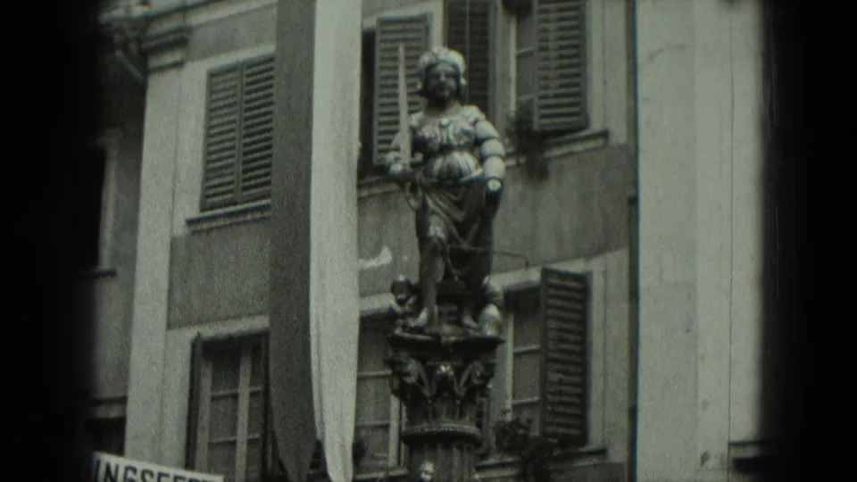 1951 Germany frame of a robed stone statue on an ornate pedestal against a shuttered building facade.