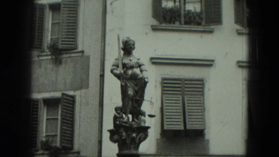 1951 West Germany home-movie frame showing a robed public statue with sword and scales against shuttered building windows.