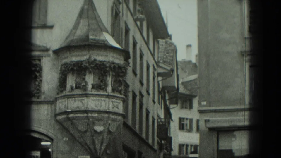 1951 West Germany home-movie frame showing a carved bay window with ivy along a narrow street of old buildings.