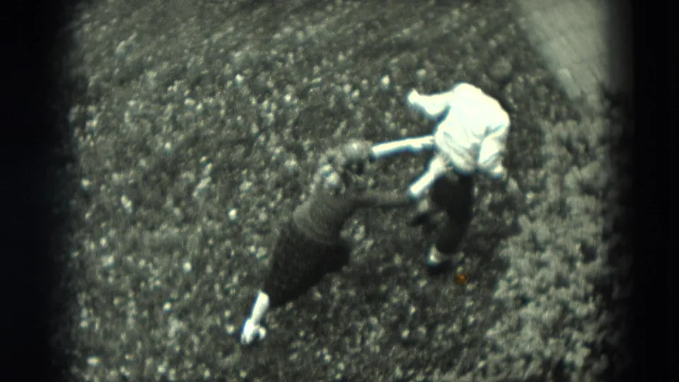 High-angle 8mm frame shows two children in a playful physical exchange on a gravel surface beside a planted edge.