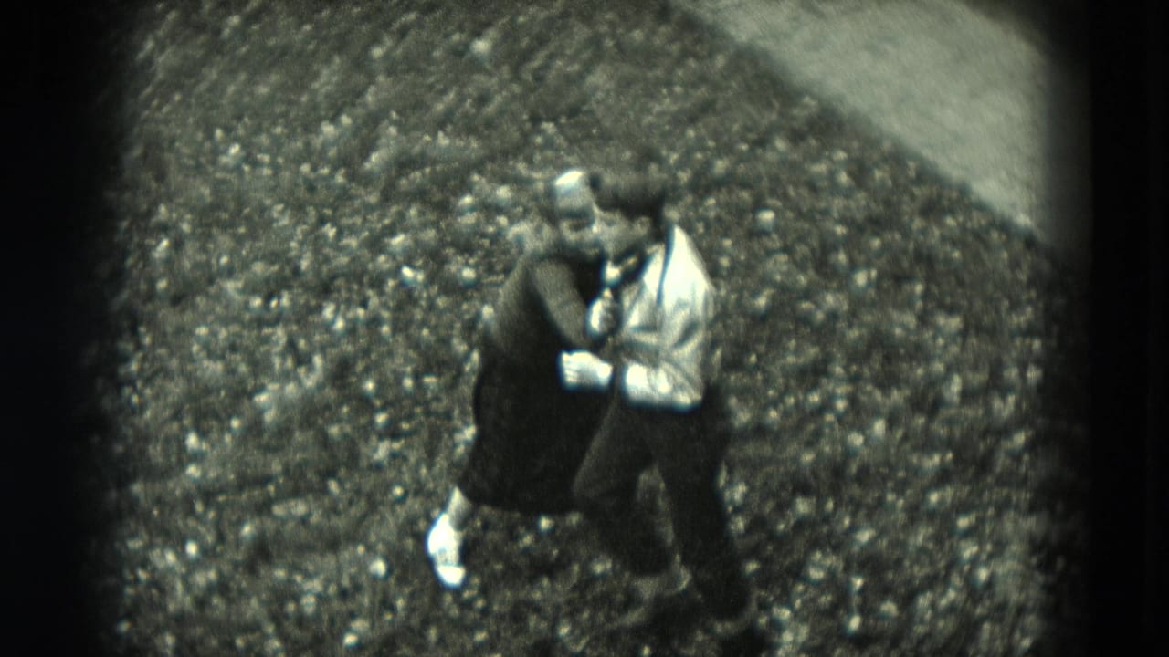 Two adults dance closely on a gravel path in a grainy black-and-white frame.