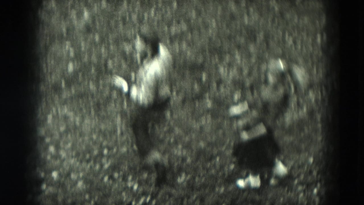 The couple appears mid-dance on an outdoor gravel path, with the man in a light shirt and the woman in a dark dress in black-and-white 8mm footage.